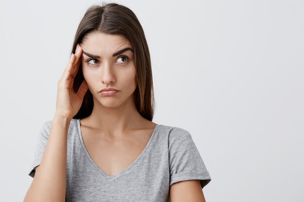 Chiuda su di giovane ragazza caucasica seria bella con capelli marroni lunghi in fronte commovente della maglietta grigia alla moda con la mano, guardando da parte con l'espressione stanca del fronte dopo la lunga giornata all'università.