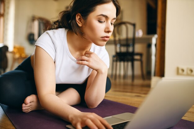Chiuda sul colpo di giovane donna concentrata che si siede sul tappetino fitness utilizzando il laptop alla ricerca di tutorial online per l'allenamento a casa, ripetendo gli esercizi dopo l'istruttore in video. Sport e gadget