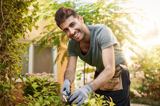 Chiuda sul ritratto all'aperto di bello allegro contadino caucasico barbuto in camicia blu e guanti sorridente, lavorando con attrezzi da giardino nella sua fattoria vicino a casa di campagna