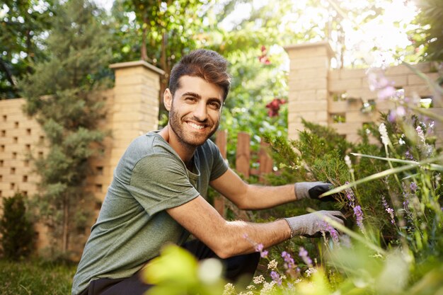 Chiuda sul ritratto del fiorista ispanico barbuto maturo attraente che sorride nella macchina fotografica, guardando sopra i fiori nel giardino vicino alla casa di campagna con l'espressione del viso felice e rilassata