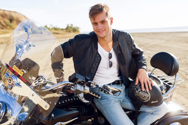 Chiuda sul ritratto del motociclista sulla strada vicino alla spiaggia. Uomo bello che si siede sulla motocicletta.