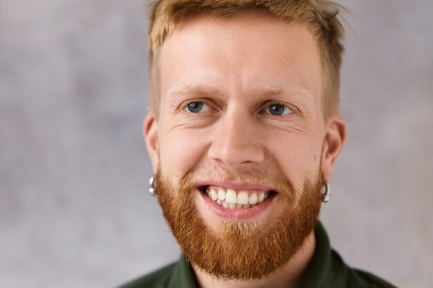 Chiuda sul ritratto del ragazzo europeo bello alla moda hipster con nuovo taglio di capelli fresco e barba folta che ride impotente guardando commedia divertente, posa isolata. Concetto di gioia, divertimento e positività