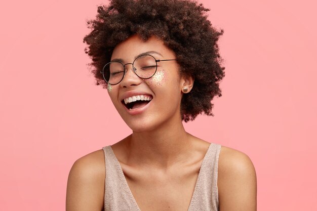 Chiuda sul ritratto della donna afroamericana felice ride di qualcosa di divertente, ha un'espressione positiva, indossa gli occhiali, ha i capelli ricci, vestito casualmente, chiude gli occhi con felicità, isolato sul rosa