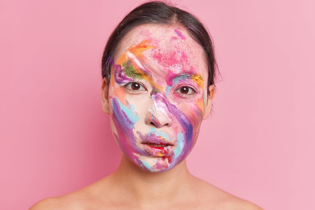 Chiuda sul ritratto della donna castana seria ha sbavature di pittura trucco creativo multicolore sul viso