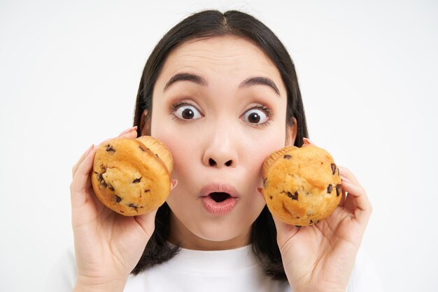 Chiuda sul ritratto della donna coreana sembra sorpreso mostra due cupcakes sfondo bianco