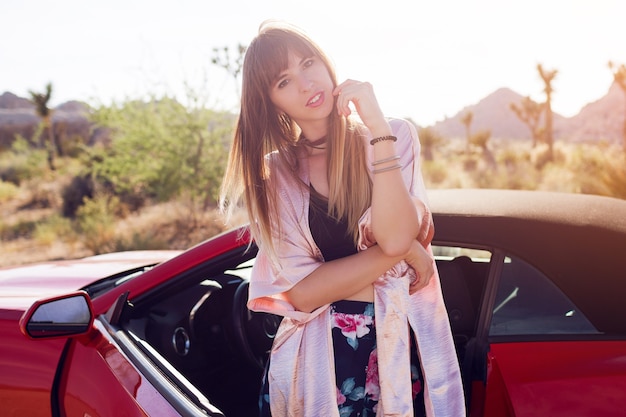 Chiuda sul ritratto della donna elegante che posa vicino alla sua automobile convertibile rossa costosa in vestito casuale. Luce della sera morbida. godendo il viaggio su strada nel parco nazionale di Joshua Tree.