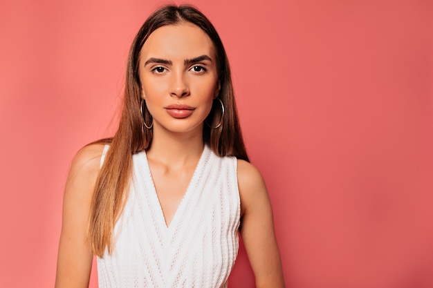 Chiuda sul ritratto della donna graziosa attraente con capelli lunghi che porta vestito bianco che posa sopra la parete rosa