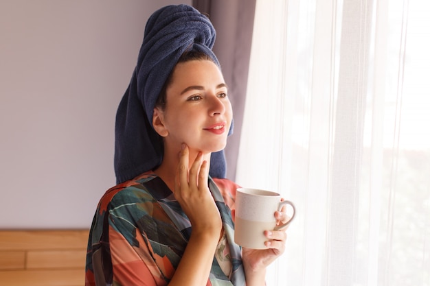 Chiuda sul ritratto della donna graziosa avvolta in asciugamano ed accappatoio che svegliano nella mattina che si siede sul letto e che beve il caffè