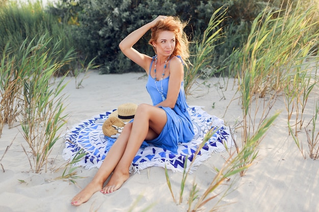 Chiuda sul ritratto della donna sorridente stupefacente della testarossa in vestito blu che si rilassa sulla spiaggia soleggiata di primavera sul tovagliolo. Cappello di paglia, eleganti bracciali e collana.