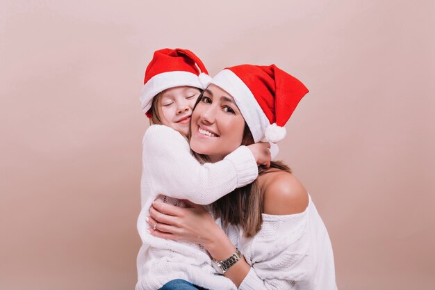 Chiuda sul ritratto della famiglia felice che indossa cappelli di Natale e maglioni bianchi, stanno abbracciando e mostrando emozioni felici reali. Muro isolato, posto per il testo