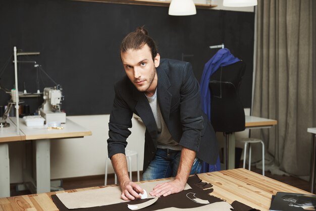 Chiuda sul ritratto dello stilista maschio caucasico moro maturo attraente che guarda in camera con l'espressione rilassata, lavorando al nuovo vestito per la raccolta dei vestiti dell'inverno.