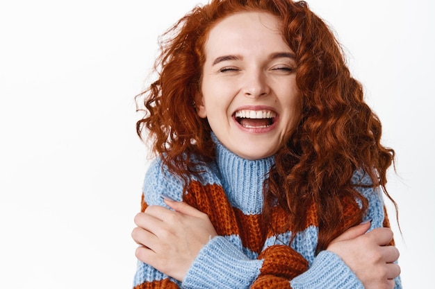 Chiuda sul ritratto di bella donna rossa felice con capelli naturali ricci, pelle liscia pallida, ridendo sincero con gli occhi chiusi e abbracciandosi su bianco