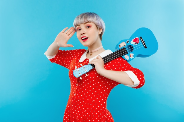 Chiuda sul ritratto di bella ragazza da bambola con brevi capelli viola chiaro che indossano le ukulele rosse della tenuta del vestito sopra la parete blu