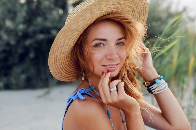 Chiuda sul ritratto di estate di bella donna allegra in cappello di paglia che si rilassa sulla spiaggia piena di sole in vacanza. Umore tropicale.