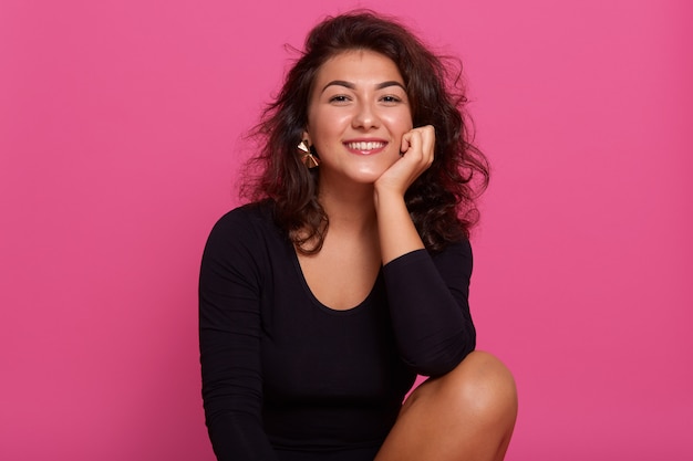 Chiuda sul ritratto di giovane bella ragazza allegra con la camicia nera vestita capelli ondulati scuri, sedersi sorridendo con i denti contro la parete rosa