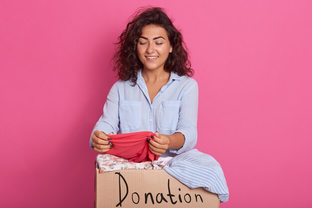 Chiuda sul ritratto di giovane donna con capelli ondulati scuri, posando vicino alla scatola di donazione dei vestiti, controllando il rosa