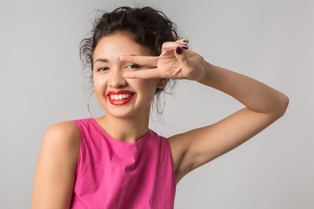 Chiuda sul ritratto di giovane donna graziosa positiva in vestito rosa, mostrando il segno di pace, felice, sorridente, stile estivo, rossetto rosso, tendenza della moda, civettuolo, asiatico, razza mista, isolato