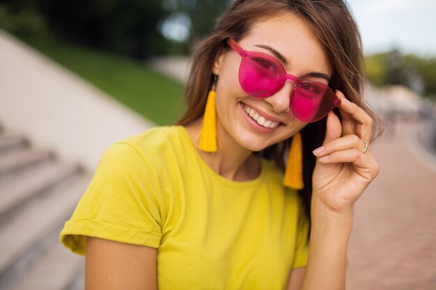 Chiuda sul ritratto di giovane donna sorridente attraente divertendosi nel parco cittadino, positivo, felice, indossa un top giallo, orecchini, occhiali da sole rosa, tendenza moda stile estivo, accessori alla moda, colorati