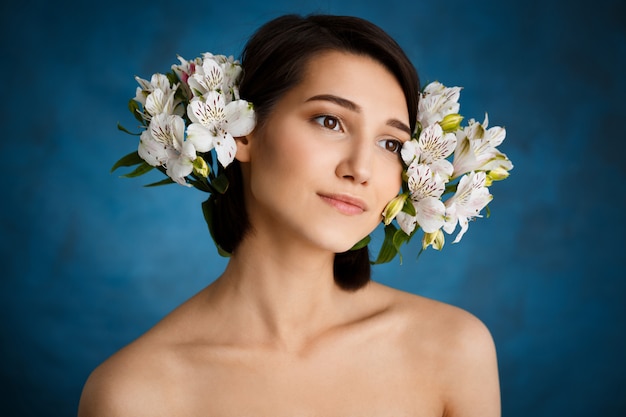 Chiuda sul ritratto di giovane donna tenera con i fiori bianchi sopra la parete blu