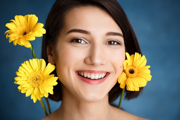 Chiuda sul ritratto di giovane donna tenera con i fiori gialli sopra la parete blu