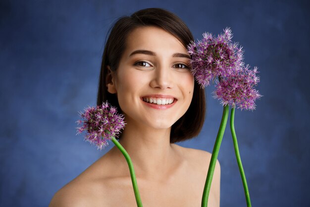 Chiuda sul ritratto di giovane donna tenera con i fiori lilla sopra la parete blu