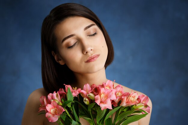 Chiuda sul ritratto di giovane donna tenera con i fiori rosa sopra la parete blu
