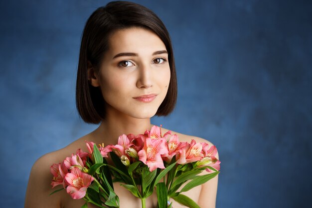 Chiuda sul ritratto di giovane donna tenera con i fiori rosa sopra la parete blu