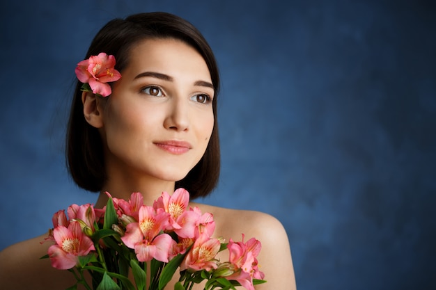 Chiuda sul ritratto di giovane donna tenera con i fiori rosa sopra la parete blu