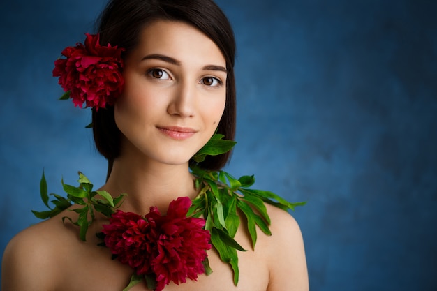 Chiuda sul ritratto di giovane donna tenera con i fiori rossi sopra la parete blu