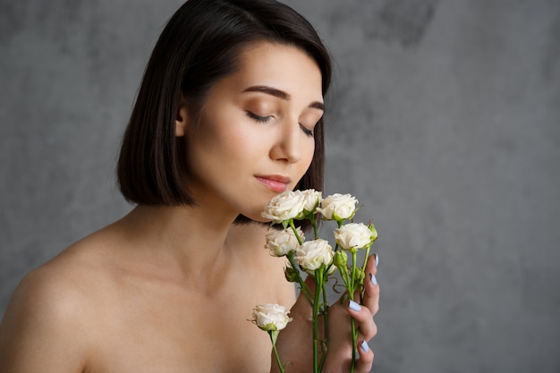 Chiuda sul ritratto di giovane donna tenera con i fiori sopra la parete grigia