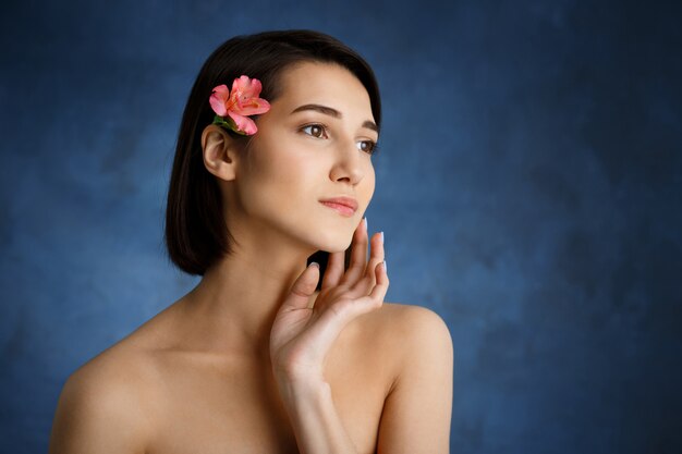 Chiuda sul ritratto di giovane donna tenera con il fiore rosa in capelli sopra la parete blu