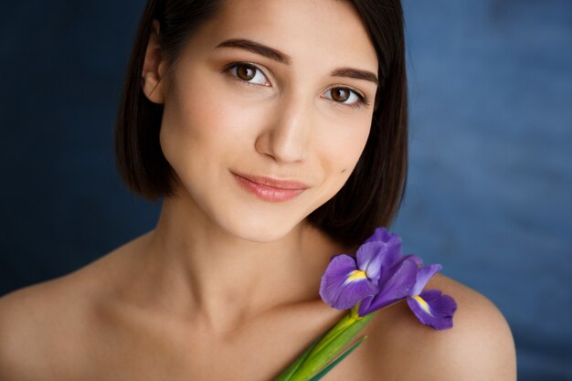 Chiuda sul ritratto di giovane donna tenera con il fiore viola sopra la parete blu