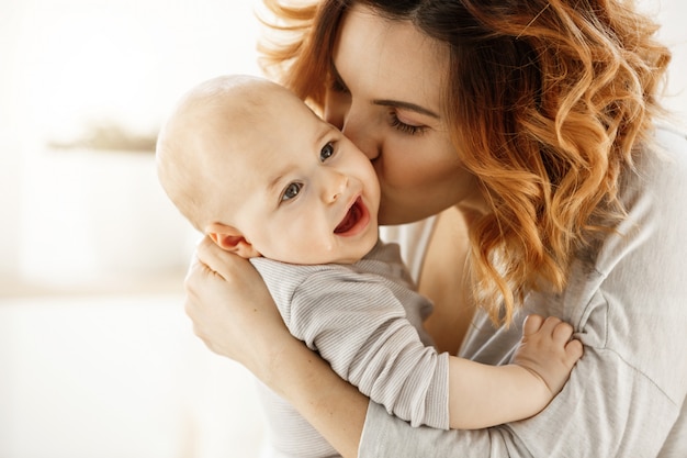 Chiuda sul ritratto di giovane madre attraente che bacia il suo bambino gridante. Kid guardando da parte con l'espressione del viso spaventato. Concetto di famiglia.