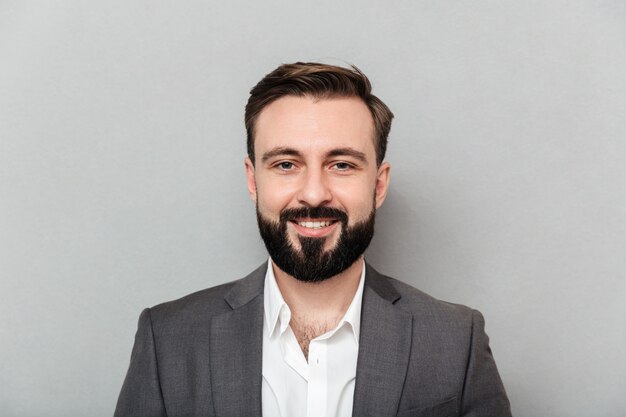 Chiuda sul ritratto di giovane uomo barbuto in camicia e rivestimento bianchi che posano sulla macchina fotografica con il vasto sorriso, isolato sopra grey