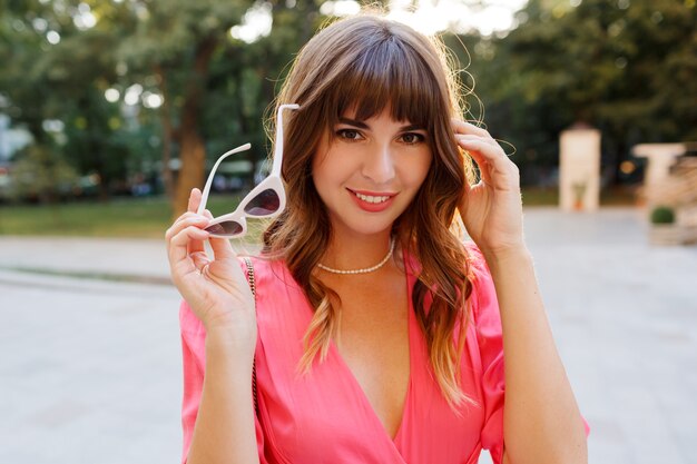 Chiuda sul ritratto di s di donna alla moda elegante con i capelli ondulati in abito rosa romantico in posa nel centro della città, i colori del tramonto.