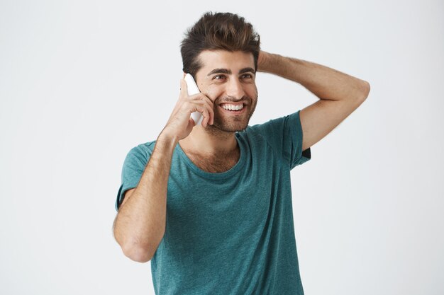Chiuda sul ritratto isolato del maschio spagnolo sorridente in maglietta d'avanguardia, sorridendo e tenendo la sua testa, mentre parla al telefono con il migliore amico.