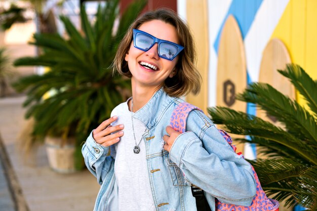 Chiuda sul ritratto positivo della donna alla moda hipster in posa davanti al surf spot, abbigliamento alla moda giovanile, occhiali da sole blu, giacca di jeans e zaino, palme intorno.