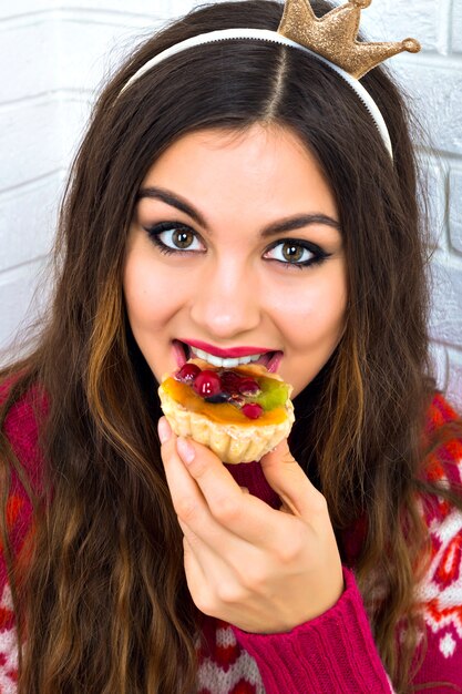 Chiuda sul ritratto urbano della donna hipster brunet divertendosi e impazzendo alla festa in maschera, indossando un maglione accogliente alla moda e una corona del viso, mangiando una torta gustosa dolce ai frutti di bosco, luminosa, positiva, felice.