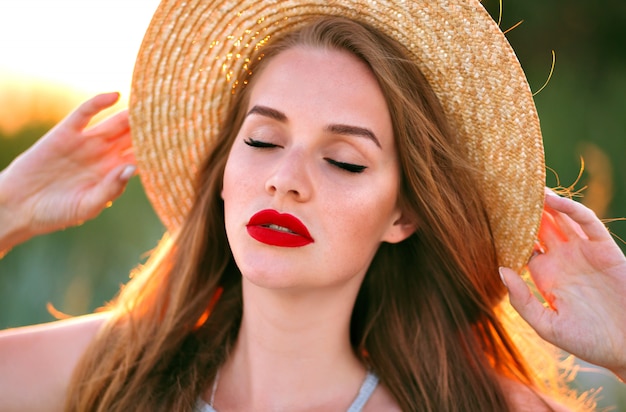 Chiuda sul tenero ritratto della donna sensuale di bellezza in posa sul campo, stile vintage, indossando il cappello alla moda di paglia, trucco di bellezza della natura, viso lentigginoso e labbra carnose rosse.