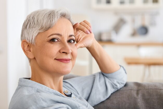 Chiuda sull'immagine della donna anziana elegante alla moda con taglio di capelli alla moda e make up rilassante al chiuso, comodamente seduto sul divano con la mano sul viso, guardando con un sorriso spensierato