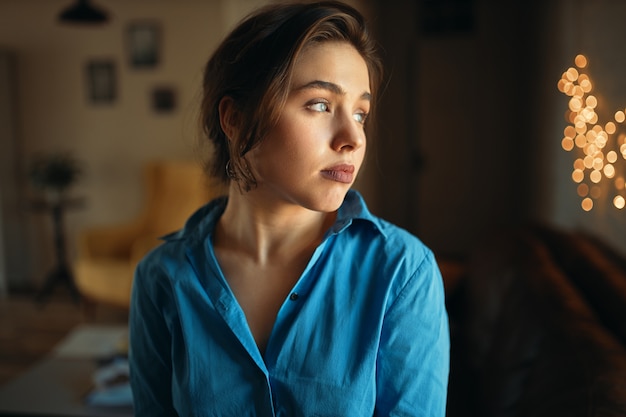 Chiuda sull'immagine della ragazza triste dell'allievo in camicia blu che distoglie lo sguardo con l'espressione facciale infelice, annoiata come deve rimanere a casa e prepararsi per l'esame. Bella giovane donna in posa all'interno