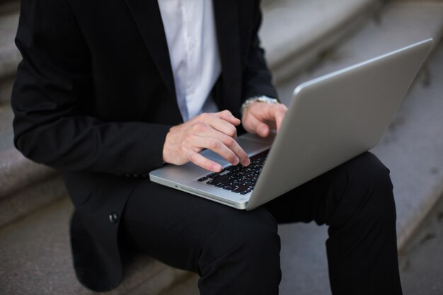 Chiuda sull'uomo d'affari nel classico vestito nero che lavora sul computer portatile all'aperto