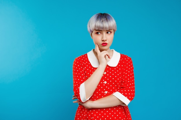 Chiuda sulla bella ragazza da bambola del ritratto con i capelli corti viola chiaro che portano il vestito rosso sopra la parete blu