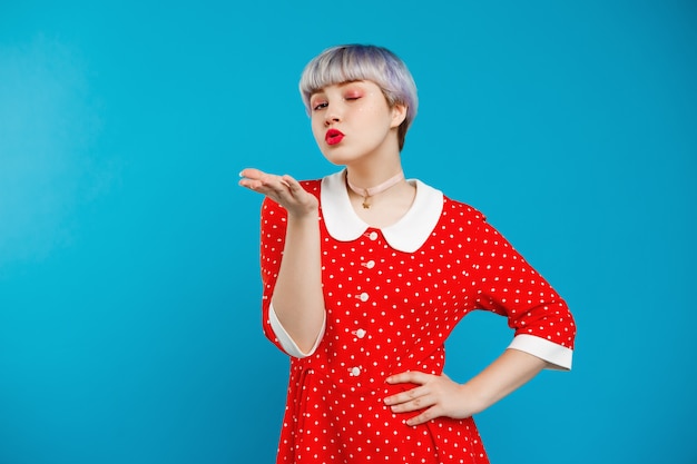 Chiuda sulla bella ragazza da bambola del ritratto con i capelli viola chiaro corti che porta il vestito rosso che invia il bacio sopra la parete blu