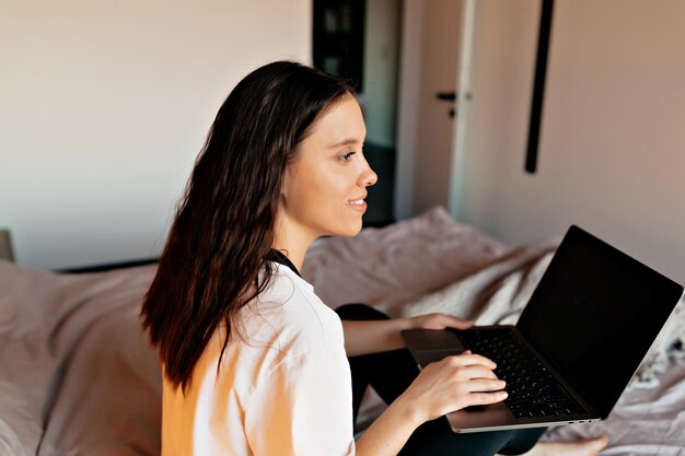 Chiuda sulla foto dell'interno della ragazza dai capelli scuri in camicia bianca sta lavorando a casa con il computer portatile a letto