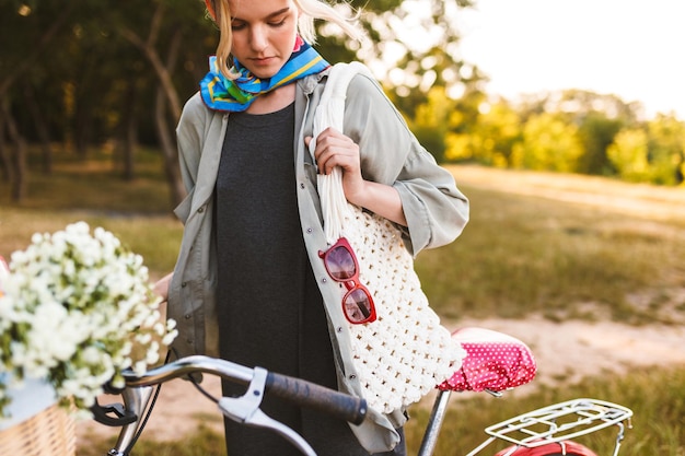 Chiuda sulla ragazza con la borsa bianca in piedi con la bicicletta e fiori di campo nel cesto nel parco