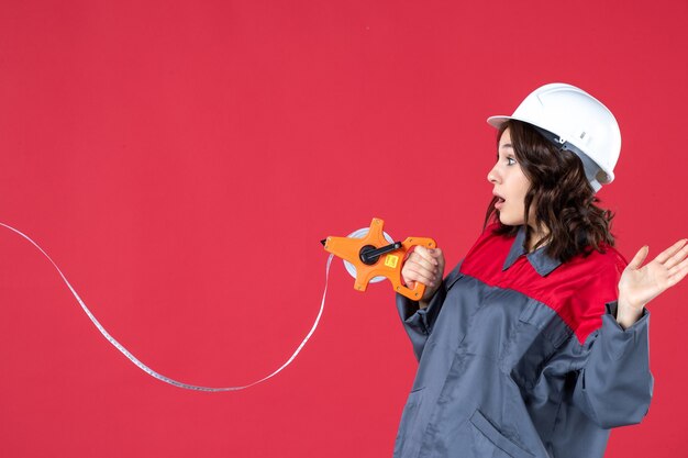 Chiuda sulla vista dell'architetto femminile scioccato in uniforme con nastro di misurazione di apertura del cappello duro sulla parete rossa isolata