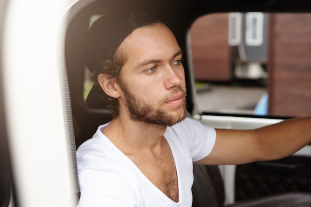 Chiuda sulla vista di giovane uomo barbuto attraente nello snapback che si siede dentro la cabina della sua jeep bianca