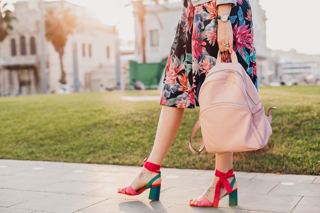 Chiudere i dettagli delle gambe in sandali rosa della donna alla moda che cammina nella strada della città in gonna colorata stampata, tenendo lo zaino in pelle rosa