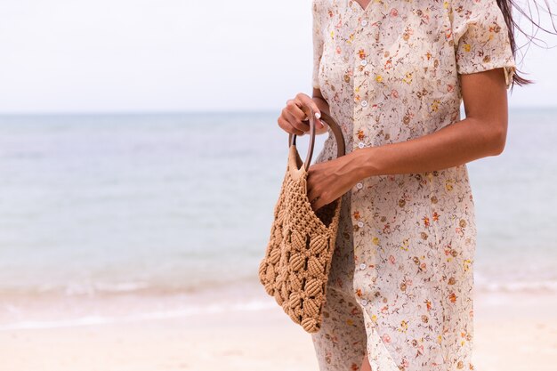 Chiudere il colpo di donna in abito volante estivo leggero che tiene borsa lavorata a maglia sulla spiaggia, mare sullo sfondo.